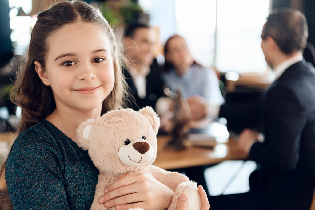 a little girl holding a teddybear with parents talking to lawyer in the background
